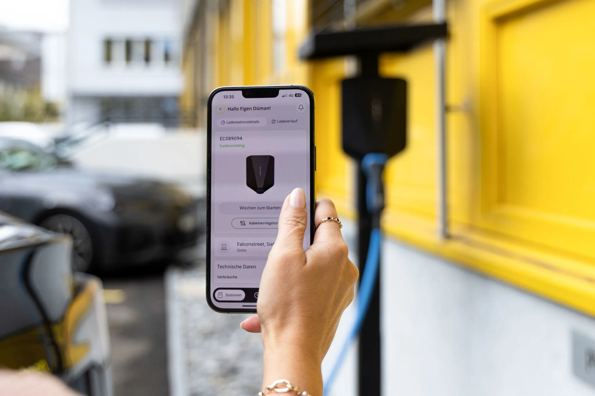 Person using a smartphone to manage charging; wallbox and yellow building in background